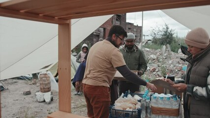 Medium shot of team of volunteers providing water, food and warm clothes to diverse people such as homeless and refugees living at tent city in poor conditions