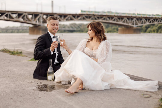 Photo of young beautiful couple, bride and groom in wedding dress are sitting on bank of wide river with glasses of wine and champagne. wedding day, happy moments.