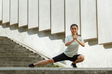 Attractive Asian lady in sportswear and sneakers does side dynamic lunges near underground crossing steps and street wall - 462884011