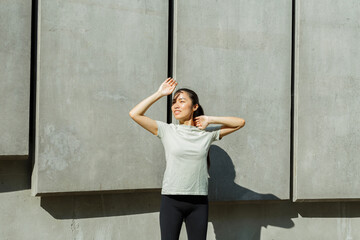 Happy young Asian sportswoman covers eyes from sun with palm standing near wall with large concrete plates on sunny city street - 462884019