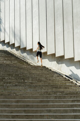 Slim woman with flying hair in tracksuit runs up large stone steps past wall with concrete plates on city street backside view