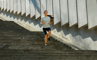 Asian woman in tracksuit runs down underground crossing large stone stairs past wall with panels training on city street in summer morning