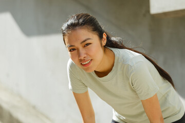 Tired asian woman with ponytail in stylish tracksuit bends leaning on knees resting at training near stone steps and concrete wall on city streetcity