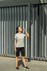 Fototapeta na wymiar Athletic Asian lady with ponytail drinks water from plastic bottle resting after training on sunny city street