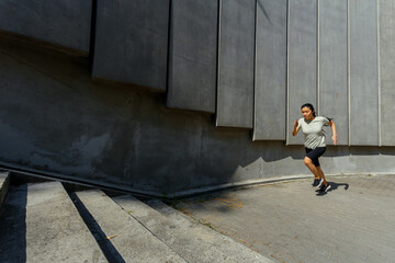 Long haired Asian woman in tracksuit runs up stone stairs past wall with concrete panels training on city street side view - 462884044