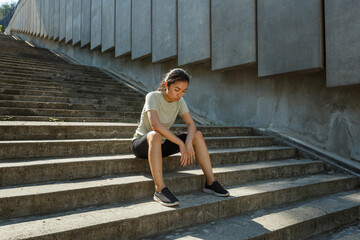 Naklejka premium Tired young Asian woman in sportswear sits on stone stairs by wall with concrete panels resting at outdoor training on street