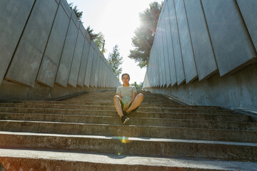Asian lady in sportswear and sneakers sits resting on underground crossing empty stone stairs...