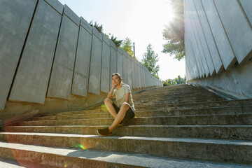 Stylish young Asian woman in tracksuit and sneakers sits resting on narrow underground crossing empty stone steps after outdoor training