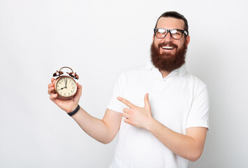Cheerful young bearded man is pointing at an alarm clock.