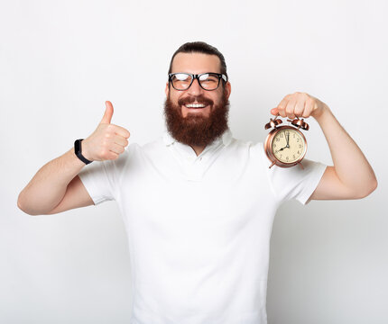 Content bearded man is showing like gesture and an alarm clock.