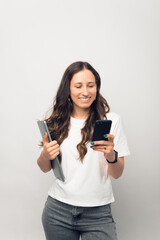 Vertical photo of a young woman is browsing on the phone while holding her laptop.