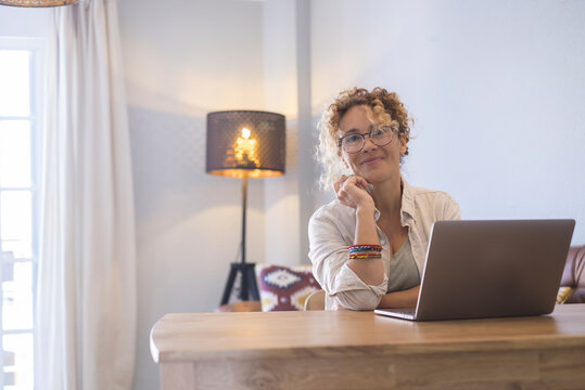 Portrait of cheerful young attractive adult businesswoman smiling and looking while work on laptop computer at home on a wood table with living room in background - modern people online job