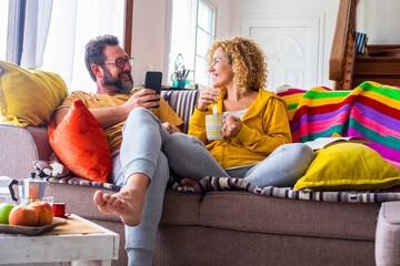 Naklejka premium Young adult happy couple enjoy leisure activity indoor at breakfast time - man use phone call and woman drinking coffee or tea - smile and happiness people sitting on the sofa enjoying home