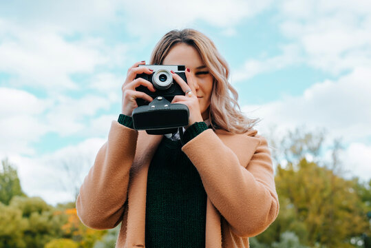 Young woman photographer with film retro camera taking picture in nature, hipster girl standing against sky and looking into vintage camera outdoors, low angle view. Hobby, tourist, travel concept