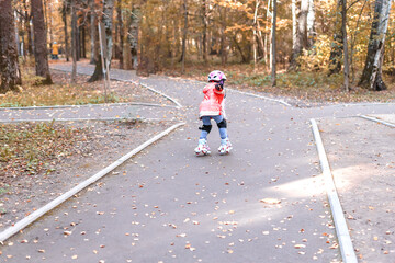 Fototapeta premium 5-year-old little girl in an orange jacket and protective clothing in an autumn park is rollerblading