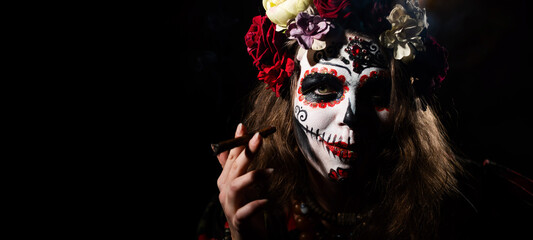 Woman in santa muerte makeup on a black background. Halloween girl smoking a cigar dressed as a...