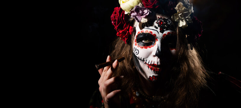 Woman in santa muerte makeup on a black background. Halloween girl smoking a cigar dressed as a traditional mexican holy death.