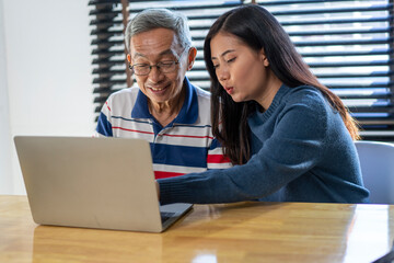 Asian granddaughter teaching old grandfather how to use the internet and reading e-mail with laptop in the living room of the house. 