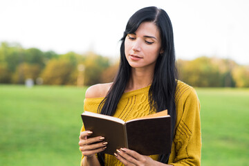 A beautiful and attractive Caucasian brunette girl in a yellow sweater is reading a book while sitting in the park.