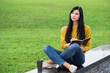 A beautiful and attractive Caucasian brunette girl in a yellow sweater writing in a notebook while sitting in the park.