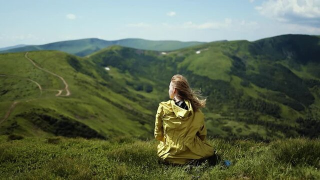Freedom blonde woman tourist in a bright yellow jacket sits on top of mountain and looking at a Mountain valley in summer sunny day. Gold hair in fresh air wind