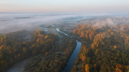 a river among fields and forests in clouds and fog with the first frosts during the golden autumn morning , top areal view from a drone