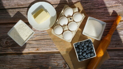 white chicken eggs wooden rolling pin bowls with sugar and flour, a bowl with fresh blueberries, slices of butter on a saucer on a wooden surface sun rays breakfast ingredients for baking a pie