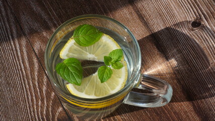 glass mug with cold lemonade, lemon slices and mint leaves on a wooden surface on a cloth napkin with a floral ornament sun rays