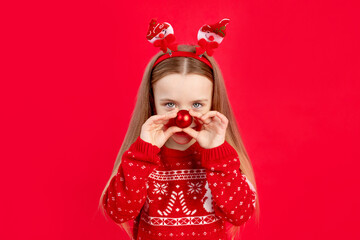 a child girl in a sweater with a ball on her nose on a red monochrome isolated background rejoices and smiles, the concept of new year and Christmas, space for text