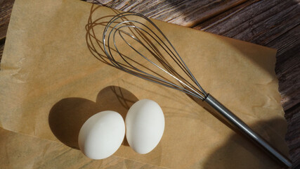 two white chicken eggs metal whisk for whipping on parchment paper on a wooden surface sun rays...