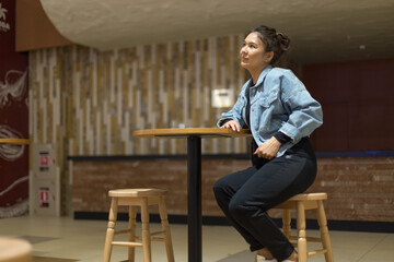girl in blue denim clothes sitting on a high bar stool in a cafe and posing for the camera, relaxed mood