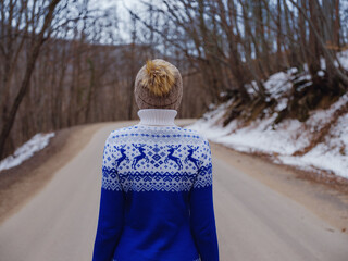 Beautiful woman standing among trees in winter forest. Wearing hat and blue sweater. Caucasian Asian female model outside in first snow