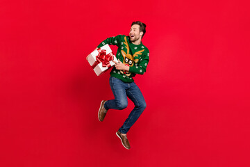 Fototapeta na wymiar Full size photo of happy smiling good mood guy in ugly sweater jump hold present box isolated on red color background