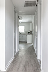 A hallway of a house leading to the kitchen with white walls, white doors and light colored laminated flooring.