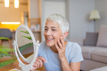 Smiling adult lady looking at herself in mirror. Beautiful senior woman checking her face skin and...