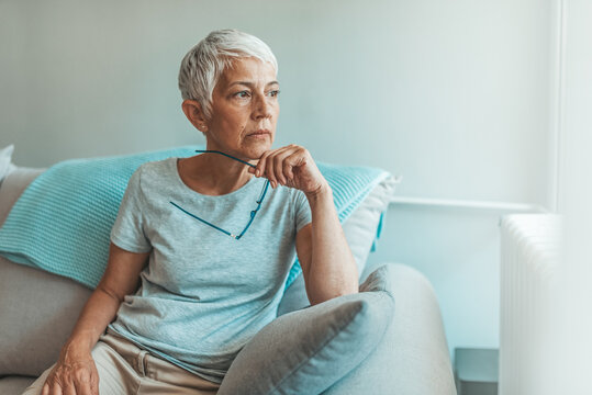 Upset depressed senior gray-haired lady touching temple, sitting on sofa, looking in the window, feeling worried and stressed about the future and thinking about own problems