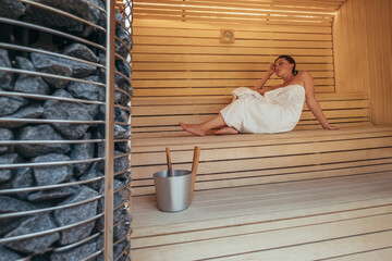 Fototapeta na wymiar Relaxing Woman wrapped white towel lying and sweating on the wooden bench in Hot Finnish sauna with hot stones and enjoying pleasant body care temperature treatment. Healthy people lifestyle concept.