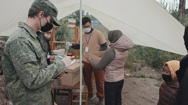 Tracking shot of team of social workers under tent providing refugees and homeless people living at immigrants camp with food and water supplies