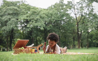 Happy little African boy lying down on mat and playing xylophone and wooden car toys at garden....