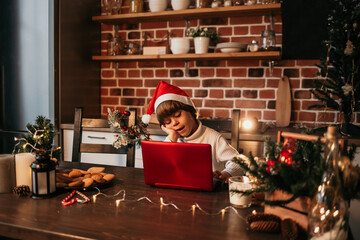 a little boy is sitting at a table in a red Christmas hat and looking at a red laptop