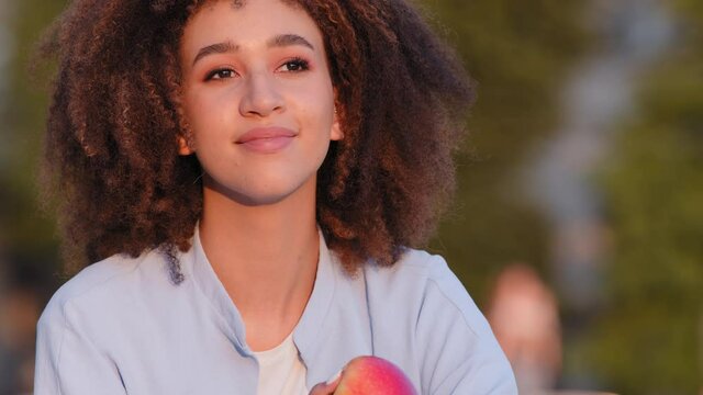 Portrait of happy african american girl afro woman ethnic mixed race lady female sitting outdoors sniffing red juicy fragrant apple applying fruit to nose enjoying scent return after covid disease