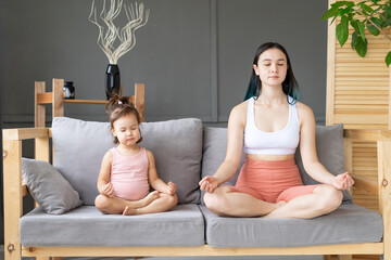 Mom and little daughter practice yoga in lotus position on sofa. Emotional connection, empathy and...