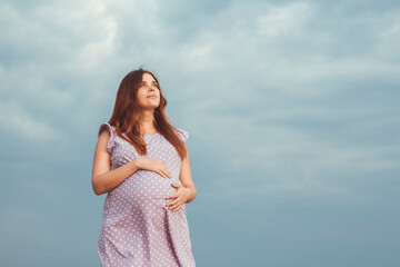 Fototapeta na wymiar red-haired pregnant woman in dress stands against background of gloomy cloudy sky caress belly and looking up to the sky with gratitude