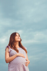 Fototapeta na wymiar red-haired pregnant woman in dress stands against background of gloomy cloudy sky caress belly and looking up to the sky with gratitude