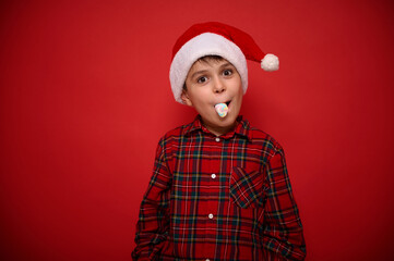 Beautiful amazing funny and cheerful preteen boy, wearing Santa hat and plaid red shirt looks at camera posing against colored background with sugary marshmallow in his mouth. Christmas concept