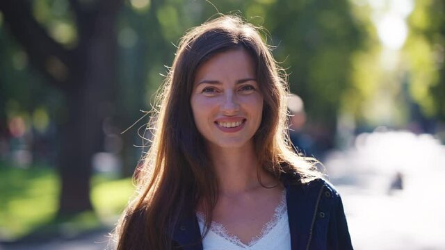 Attractive Woman Wearing Jacket Smiling At Camera In Park, Sunshine In Her Hair, Blurred People Walking On Background, Parallax Effect. Portrait Of Happy Female Outside. Concept Of Emotion