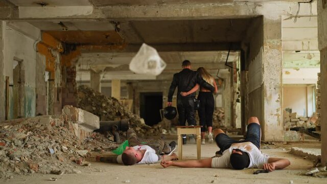 Hugging couple of man in moto costume and saved blonde woman leaves premise while dead criminals lie in abandoned building backside view slow motion