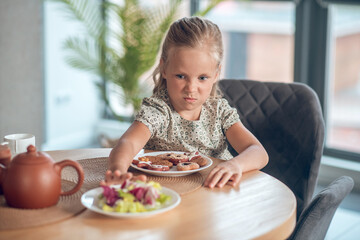 A small girl looking unpleased while looking at the food on the table