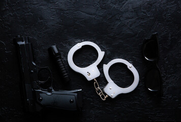 Black gun, flashlight, glasses and Police metal real handcuffs lie on the black background. Private...