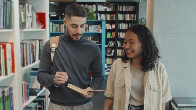 Tracking Medium Slowmo Shot Of Multi-ethnic Couple Of Young Adult University Students In Casualwear Discussing Book While Walking Along Library